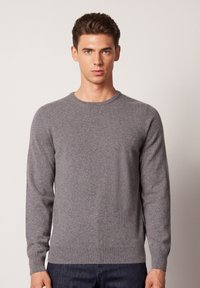 Grau gestrickter Pullover mit rundem Halsausschnitt und langen Ärmeln. Er hat eine weiche Textur und eine taillierte Passform und wird über dunklen Jeans getragen.