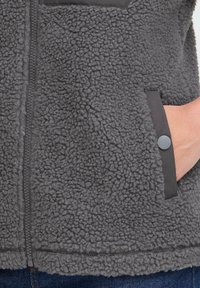 Gros plan sur une veste en polaire grise avec une fermeture éclair et une poche latérale où une main est partiellement insérée.