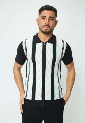 Veste zippée à manches courtes avec un col noir, présentant un motif en losanges blancs et des rayures verticales noires, fabriquée en tissu tricoté.