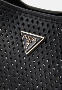 Czarny perforowany materiał z trójkątnym metalowym logo Guess USA, w którym znajduje się znak zapytania.