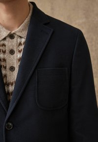 Blazer di lana blu con una tasca sul petto e revers a scollo, indossato sopra una maglietta in maglia beige con motivo e bottoni marroni.
