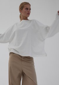 Biała, oversize'owa koszulka z długim rękawem wykonana z miękkiego materiału, zestawiona z jasnobrązowymi spodniami z bezszwowym paskiem i bocznymi kieszeniami.