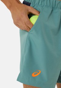 Men's teal athletic shorts with a textured finish, orange logo, and pocket holding a yellow tennis ball. Elastic waistband visible.