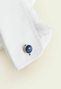 Weißer, strukturiert gewebter Hemdärmelsaum mit einem marineblauen, stoffbezogenen Manschettenknopf, der eine kleine rosa Blumenstickerei zeigt.