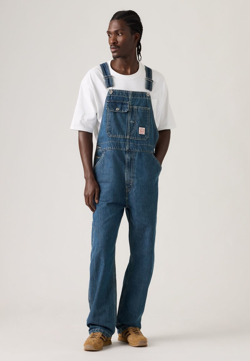 Τζιν φορμάκι με άνετη γραμμή, σκούρο μπλε χρώμα, μπροστινές τσέπες, ρυθμιζόμενες τιράντες και λογότυπο σε μπαλώμα. Συνδυασμένο με λευκό T-shirt και καφέ παπούτσια.