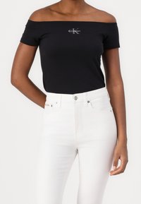 Top negro sin hombros hecho de algodón, con un pequeño logo plateado de Calvin Klein. Combinado con jeans blancos de cintura alta.