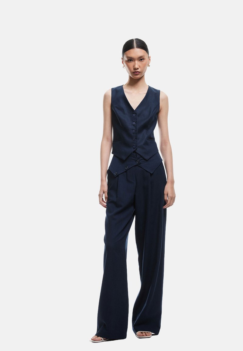 Navyblauwe asymmetrische vest met een v-hals, knopen aan de voorkant; wijde broek met plooien en zijzakken. Glad stoftextuur.