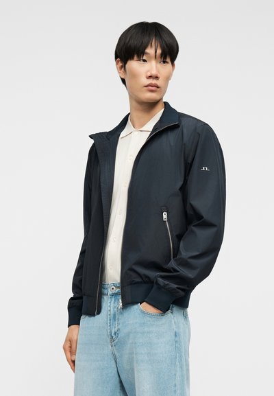 Νέος άνδρας με μπλε μπουφάν με φερμουάρ, λευκό πουκάμισο με κουμπιά και ανοιχτόχρωμο τζιν, στέκεται με το ένα χέρι στην τσέπη, κοιτάζοντας πλάγια.