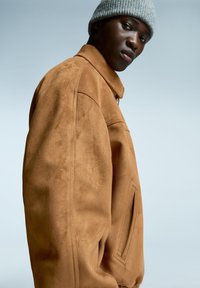 Veste en daim marron avec un col structuré, une poche latérale et une texture lisse, associée à un bonnet tricoté gris clair.