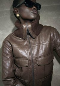 Giacca di pelle marrone con colletto alto e rigato, chiusura con zip e due tasche sul petto. Il modello indossa occhiali da sole neri e orecchini a cerchio dorati.