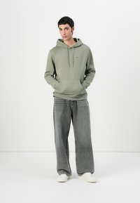 Olive green hoodie with a kangaroo pocket and drawstring hood, paired with loose gray jeans and white sneakers. Simple, casual design.