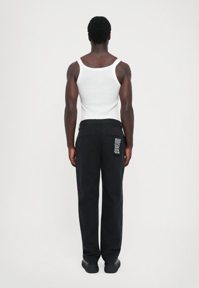 Black pants with a relaxed fit, featuring red accents and a textured patch. Worn with a white tank top, viewed from the back.