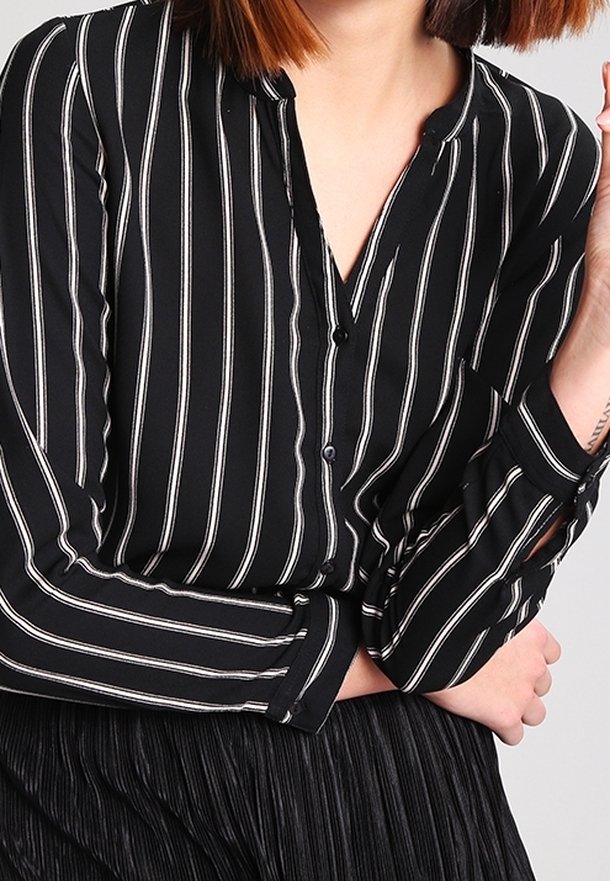 Blouse noire à rayures fines blanches verticales, sans col, à manches longues, avec des boutons au niveau du poignet.
