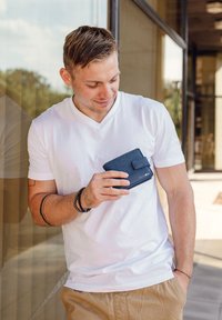 Navy blue wallet with a textured finish and magnetic closure held by a man wearing a white V-neck t-shirt, standing against a glass window.