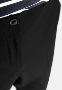 Czarne spodnie o teksturowanym wykończeniu, z zapięciem na guzik i widoczną z przodu kieszenią blisko pasa. Częściowo widoczna koszula w paski.