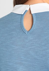 Blå stickad topp med vit krage och en nyckelhålskonstruktion på baksidan, med en texturerad finish och slät yta.