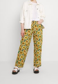 Pantalones de pierna ancha con un patrón de girasoles en amarillo y verde, combinados con una camiseta blanca y una chaqueta de mezclilla color crema. Sandalias negras.