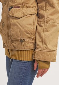 Gros plan d'une personne portant une veste matelassée marron clair avec poches à rabat, ourlet côtelé et jean bleu, la main détendue le long du corps.