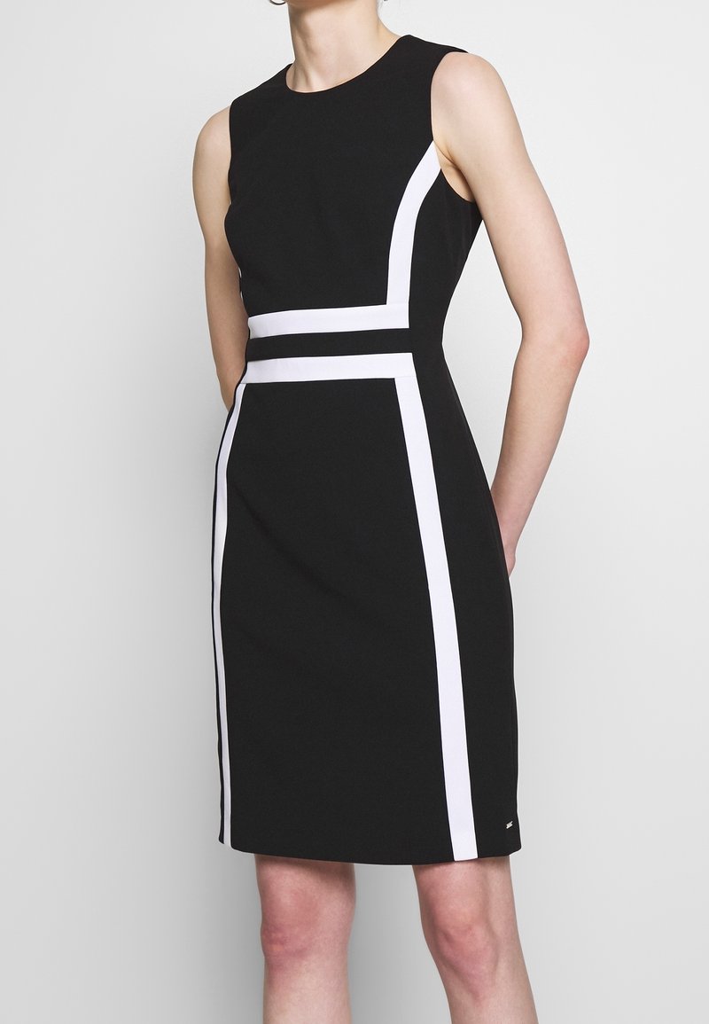 Robe noire sans manches avec des rayures blanches verticales et horizontales formant un motif géométrique, silhouette ajustée, portée par une personne debout.