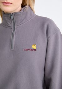 Primer plano de una persona que lleva una sudadera gris de Carhartt con cremallera de un cuarto, con el logo amarillo y rojo bordado en el pecho.