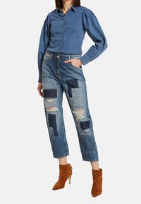 Denim-paita nappihihalla ja puffihihalla, yhdistettynä kuluneisiin farkkuihin, joissa on eri sinisten sävyjen tilkkutöitä; ruskeat nilkkurit.