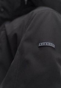 Schwarze Jacke aus strukturiertem Stoff mit einem kleinen, rechteckigen Etikett mit der Aufschrift "bugatti" am linken Ärmel.
