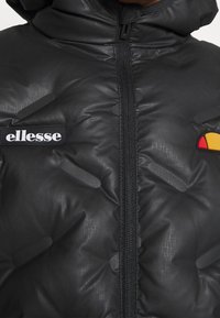 Giacca a piuma nera con finitura lucida, chiusura con zip frontale e loghi. Presenta un cappuccio, texture trapuntata e dettagli in gomma rossi e gialli.