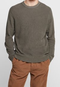 Olivgrüner gerippter Pullover mit Rundhalsausschnitt, langen Ärmeln und strukturierten Stoff. Getragen mit braunen Cordhosen mit vertikalen Streifen.