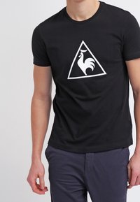 Svart bomullströja med en vit, triangulär logotyp med en stiliserad tuppdesign i mitten. Kortärmad och med rund halsringning.