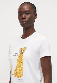 Balta kokvilnas T-kreklā ir dzeltena leoparda ilustrācija, kas valkā brilles, ar tekstu "SLIKTAS VIBRĀCIJAS" un "NEIET AR MANU LŪPU KRĀSU."