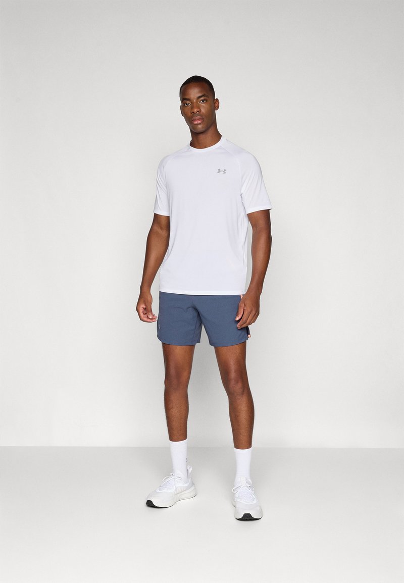 Weißes sportliches Kurzarmshirt mit grauem Logo, kombiniert mit marineblauen Shorts. Das Modell trägt weiße Sneakers und weiße Knöchelsocken und steht vor einem grauen Hintergrund.