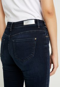 Jeans de mezclilla azul oscuro con cinco bolsillos, remaches dorados y una etiqueta reflectante. Corte ajustado con un detalle de costura sutil.