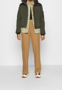 Persona che indossa una giacca imbottita verde, una camicia a quadri verde e beige, pantaloni a coste color cammello e sneakers bianche con accenti arancioni e neri.