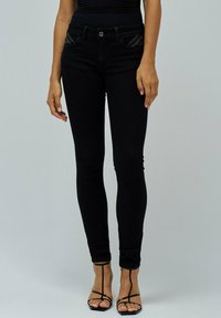 Mujer con vaqueros ajustados negros con bolsillos frontales con tachuelas y sandalias de tacón alto de tiras negras frente a un fondo liso.