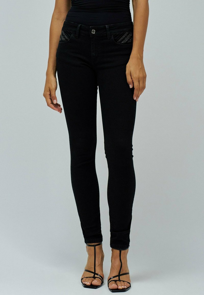 Mujer con vaqueros ajustados negros con bolsillos frontales con tachuelas y sandalias de tacón alto de tiras negras frente a un fondo liso.