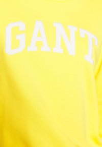 Žlutá mikina s bílým vyšívaným nápisem "GANT". Materiál je měkký a strukturovaný, s designem crew neck.