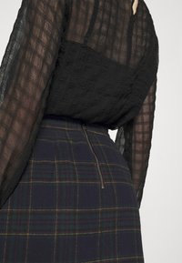 Svart strukturerad blus med långa ärmar, kombinerad med en marinblå rutig kjol med gröna och röda linjer samt en sidodragkedjedetalj.