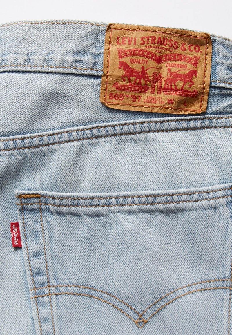 Jeans de mezclilla azul claro con una etiqueta de cuero marrón que presenta texto rojo y un diseño de caballos. La costura es de un color dorado contrastante.
