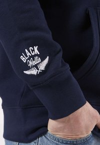 Sweat à capuche bleu marine avec le texte brodé en blanc "BLACK" et "Wells" ainsi qu'un motif de fougère sur la manche gauche. Tissu en coton doux.