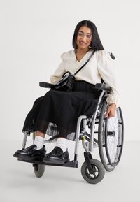 Utilisateur de fauteuil roulant assis, portant un chemisier crème et une jupe plissée noire. Chaussures noires avec des chaussettes blanches. Sac à main noir sur l'épaule.
