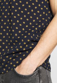 Brazo masculino de tono de piel clara que lleva una camisa de color azul oscuro con pequeños patrones de hojas amarillas, mano parcialmente en el bolsillo delantero de unos jeans negros descoloridos.