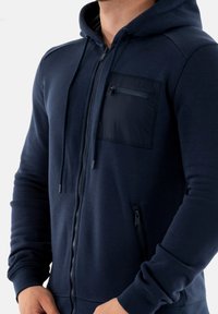 Felpa con zip di colore blu navy, realizzata in un tessuto testurizzato, con tasca frontale sul petto e tasca laterale con zip a contrasto. Presenta lacci e polsini a costine.