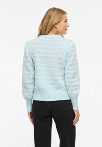 Femme portant un pull en tricot bleu clair orné de pompons blancs, associé à un pantalon noir, debout devant un fond blanc uni.