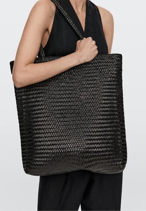 Person trägt schwarzen ärmellosen Oberteil und Hose und hält eine große schwarze gewebte Tragetasche über der Schulter vor einfachem Hintergrund.