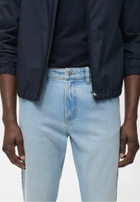 Laivaston vetoketjullinen takki mustan paidan päällä, yhdistetty vaaleansinisiin farkkuihin. Pehmeä farkkukangas, näkyvät saumat ja metallinen nappikoriste.