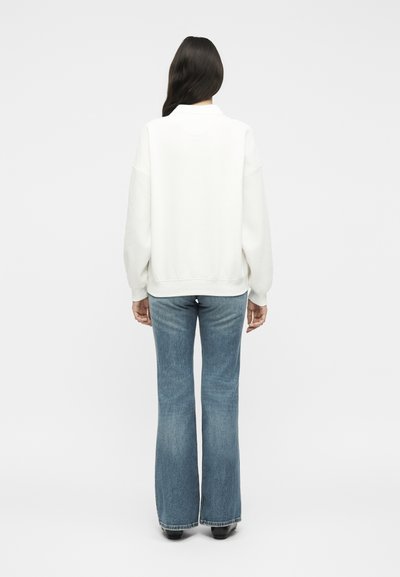 Personne aux longs cheveux foncés portant un sweat-shirt blanc et un jean évasé bleu, debout de dos devant un fond blanc uni.