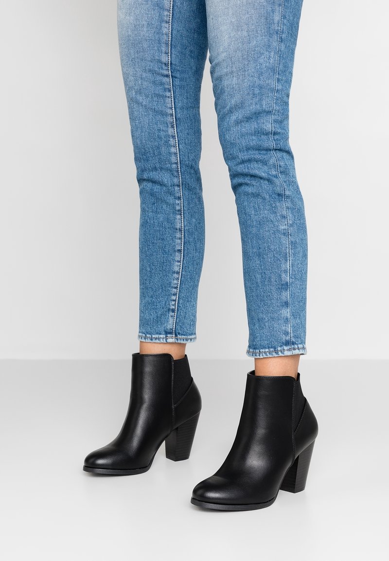 Ben klædt i tætsiddende blå jeans og sorte læder ankelsko med kraftige hæle står mod en ensfarvet lys baggrund.