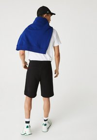 T-shirt blanc, shorts noirs et un pull bleu drapé sur les épaules. Porte une casquette noire, des baskets blanches et vertes avec des chaussettes montantes.