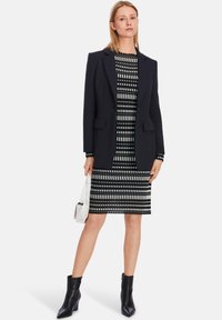 Schwarze Blazer über einem gestreiften schwarz-weißen Kleid mit knielangem Saum. Durchsichtige Stoffschuhe mit Blockabsatz, hält eine kleine weiße Tasche.