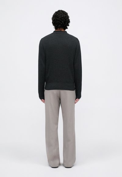 Uomo in piedi, di spalle, che indossa un maglione a coste nero e pantaloni grigi chiari larghi, su uno sfondo bianco semplice.